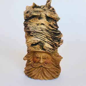 May include: A carved wooden sculpture of a bearded man with a hat. The sculpture is made of light brown wood with a textured, layered appearance. The man's face has detailed features, including a long beard and a stern expression.