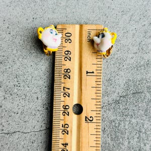 Puede incluir: Un par de pendientes de clip con un diseño de tetera de dibujos animados. Los pendientes son blancos con detalles amarillos y clips dorados. Los pendientes miden aproximadamente 2,5 cm de largo.