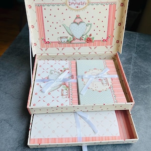 May include: A decorative gift box with a floral and heart pattern, featuring a teapot illustration. Inside are two bound notebooks and stationery, tied with white ribbons. The box is open, revealing the contents, with a drawer below.