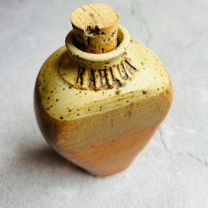 May include: A ceramic bottle with a cork stopper. The bottle is decorated with brown and beige glaze and has the word "Kahlua" written on the side.