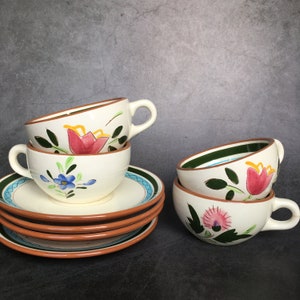 Puede incluir: Un juego de tazas y platillos de cerámica blanca. Las tazas y platillos tienen un borde marrón y están decorados con flores pintadas a mano en rosa, azul y verde. Los platillos tienen un borde azul y verde.