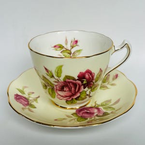 May include: A vintage teacup and saucer set with a floral design. The cup and saucer are white with a yellow background and feature pink roses with green leaves. The set has a gold trim around the edges.