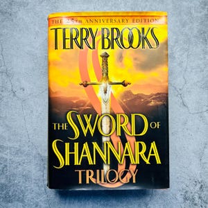 Puede incluir: Portada de "The Sword of Shannara Trilogy" de Terry Brooks, edición del 25 aniversario. Presenta una espada dorada sobre un fondo naranja y amarillo, con el título en letras amarillas.