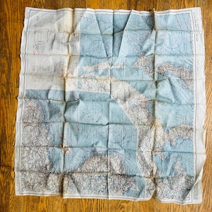 May include: A vintage map with a blue and white color scheme, featuring detailed geographical lines and markings. The map is folded and appears to be made of paper, with some areas showing signs of wear. The map is laid out on a wooden surface.