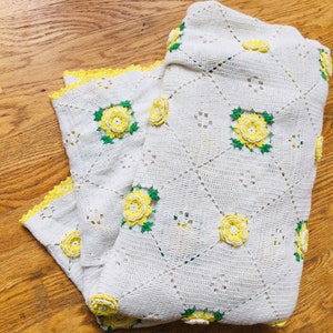May include: A white crocheted blanket with yellow rose appliqués and green leaves. The blanket has a diamond pattern and a yellow scalloped edge. The handmade blanket is folded on a wooden surface.
