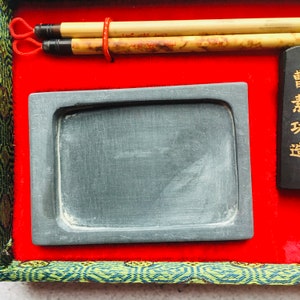 May include: A calligraphy set featuring two bamboo brushes, a rectangular ink stone, and a black ink stick with gold text. The set is presented on a red velvet surface, framed by green and gold patterned fabric.