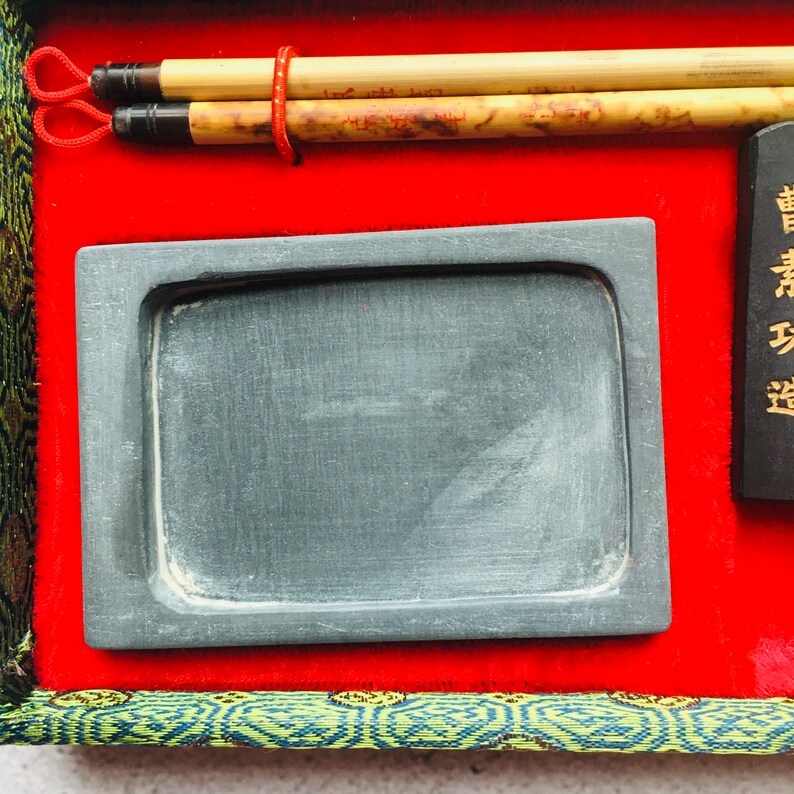 May include: A calligraphy set featuring two bamboo brushes, a rectangular ink stone, and a black ink stick with gold text. The set is presented on a red velvet surface, framed by green and gold patterned fabric.