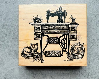 May include: A wooden stamp featuring a detailed illustration of a vintage sewing machine. A cat sits near the machine, with a basket of yarn and sewing notions. The design is in black ink on a light wood block.