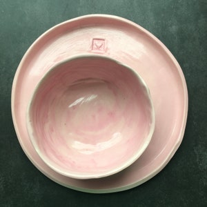Può includere: Un set di ciotola e piatto in ceramica rosa. La ciotola è più piccola e si trova all'interno del piatto più grande. Entrambi i pezzi hanno un design marmoreo sottile.