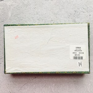 May include: A rectangular box with a white interior and a green and gold patterned border. A sticker on the box reads "URBAN OUTFITTERS" and includes a barcode and other details. The number "20" is written in red.