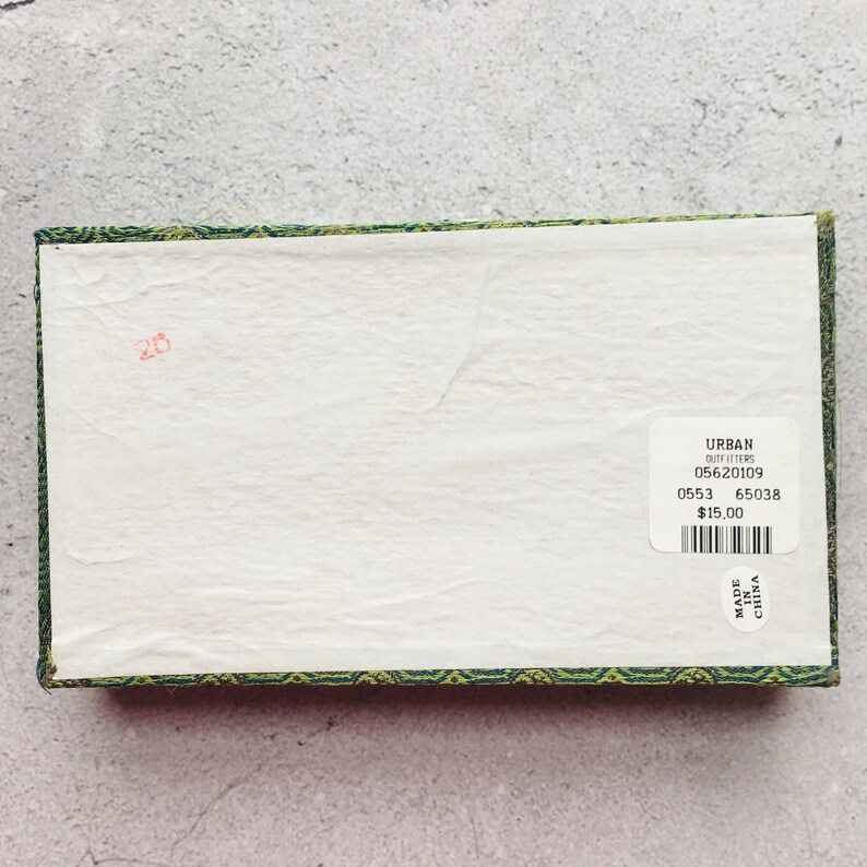 May include: A rectangular box with a white interior and a green and gold patterned border. A sticker on the box reads "URBAN OUTFITTERS" and includes a barcode and other details. The number "20" is written in red.