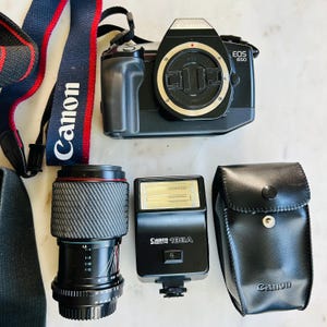 May include: A black Canon EOS 650 camera with a lens, flash, and carrying case. The camera has a black body with a silver lens mount. The lens is black with red accents. The flash is black with the text "Canon 188A". A navy blue and red Canon strap is also included.
