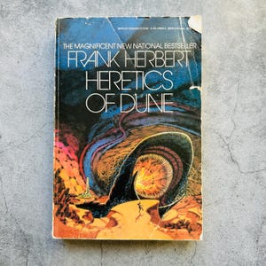 May include: A vintage paperback book titled "Heretics of Dune" by Frank Herbert. The cover features a vibrant illustration with a large, dark creature emerging from a desert landscape. The book is a national bestseller.