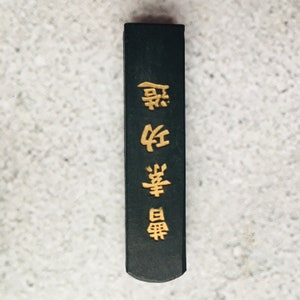 May include: A rectangular, black ink stick with gold Chinese characters. The ink stick has a slightly rounded end and is set against a light gray background. The gold characters are vertically aligned.