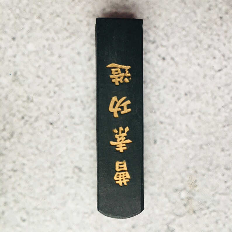 May include: A rectangular, black ink stick with gold Chinese characters. The ink stick has a slightly rounded end and is set against a light gray background. The gold characters are vertically aligned.