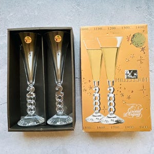 May include: Two clear champagne flutes with a spiral stem design, each featuring the number "2000". They are displayed in a black box, alongside a vintage box with the text "Millennium" and "Cristal d'Arques".