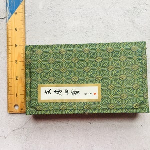 May include: A small, rectangular green box with a repeating geometric pattern. A white label with black Japanese characters is affixed to the front. A wooden ruler is placed next to the box, measuring in inches.