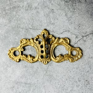 May include: Ornate gold-colored furniture hardware with a symmetrical design. The piece features two circular openings and a central decorative element with intricate detailing. The hardware is likely made of metal and has a vintage or antique aesthetic.