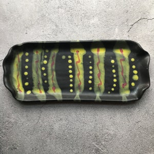Puede incluir: Bandeja de servir de cerámica negra con un diseño abstracto ondulado en amarillo y rojo. La bandeja tiene una forma rectangular con bordes redondeados.
