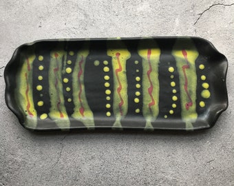 May include: Black ceramic serving tray with a wavy, abstract design in yellow and red. The tray has a rectangular shape with rounded edges.