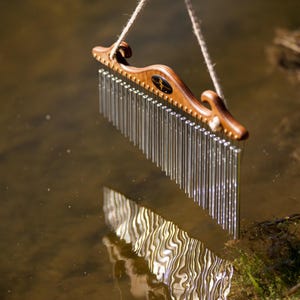 Puede incluir: Un carillón de viento de madera con una parte superior curvada y numerosos tubos de metal plateado colgando debajo. El carillón está suspendido por una cuerda, con su reflejo visible en el agua. La madera tiene un color oscuro y rico.