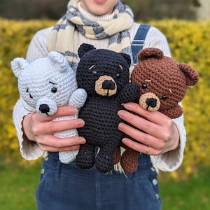Peut inclure: Une personne tenant trois animaux en peluche crochetés : un ours polaire blanc, un ours noir et un ours brun. Les ours ont des yeux noirs et des nez en bouton.