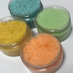 Peut inclure: Quatre petits pots en plastique transparent remplis de gommages pour le corps colorés. Les gommages sont dans les tons jaune, orange, vert et bleu. Le gommage bleu a de petits points noirs. Les pots sont disposés en gros plan.