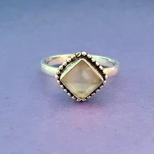 May include: A silver ring with a white gemstone set in a square bezel. The bezel has a beaded edge.