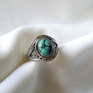 Peut inclure: Une bague en argent avec une pierre turquoise sertie dans une bélière décorative. La bague présente un design détaillé avec une bordure en forme de corde.