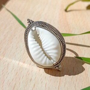 May include: A silver ring with a white cowrie shell set in a decorative border. The ring is on a wooden surface.