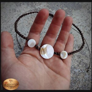 May include: A brown cord necklace with three white and brown shell pendants. The necklace is displayed on a hand.