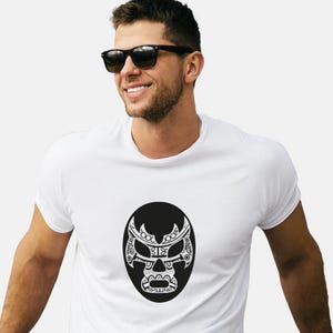 May include: White t-shirt featuring a black and white graphic of a luchador mask. The mask design is detailed with intricate patterns and is set against a black oval. The shirt is a classic crew neck style.