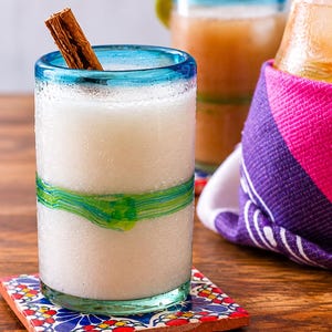 Puede incluir: Un vaso transparente con borde azul y un detalle de vidrio verde en espiral alrededor del centro, lleno de un l&iacute;quido blanco opaco y una rama de canela marr&oacute;n. El vaso descansa sobre un posavasos cuadrado con un patr&oacute;n floral colorido.