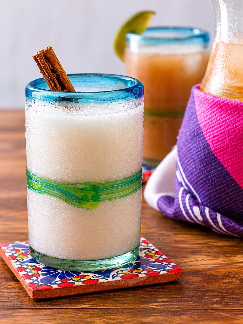 Puede incluir: Un vaso transparente con borde azul y un detalle de vidrio verde en espiral alrededor del centro, lleno de un l&iacute;quido blanco opaco y una rama de canela marr&oacute;n. El vaso descansa sobre un posavasos cuadrado con un patr&oacute;n floral colorido.