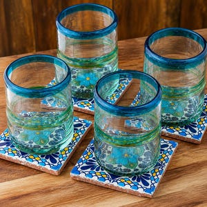 May include: Four clear glass tumblers with thick teal rims, each resting on a square ceramic coaster featuring a vibrant blue, yellow, and white floral pattern. The glasses have a slightly textured, hand-blown appearance with subtle green tints.