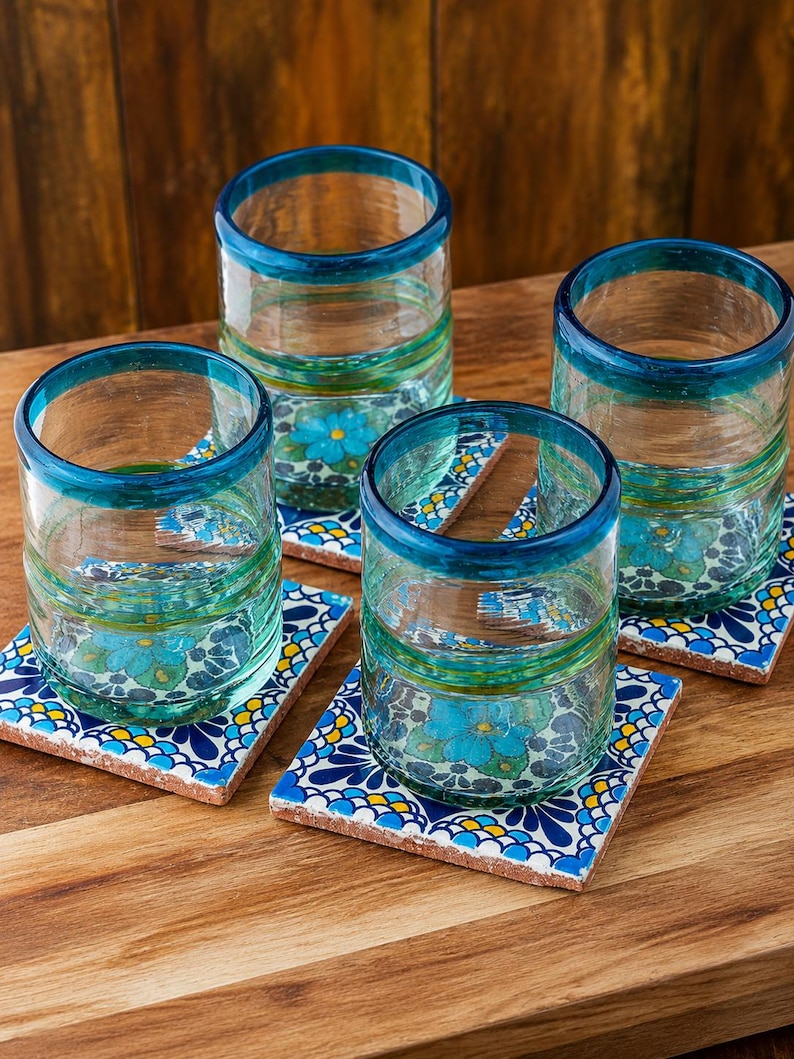 May include: Four clear glass tumblers with thick teal rims, each resting on a square ceramic coaster featuring a vibrant blue, yellow, and white floral pattern. The glasses have a slightly textured, hand-blown appearance with subtle green tints.