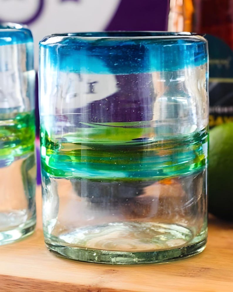 May include: A clear glass tumbler featuring a thick blue rim and horizontal bands of green and teal glass swirls. The cylindrical vessel has a slightly uneven, hand-blown texture with visible air bubbles throughout the transparent glass structure.