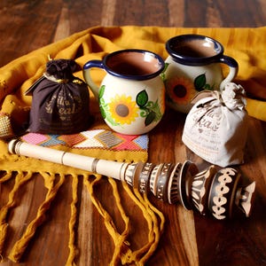 Könnte beinhalten: Zwei traditionelle mexikanische Tassen mit blauen Rändern und Sonnenblumendesigns, ein hölzerner Molinillo-Schneebesen und zwei Beutel mit Schokoladenpulver. Die Beutel sind mit "La Real" und "Chocolate para preparar" auf Spanisch beschriftet.