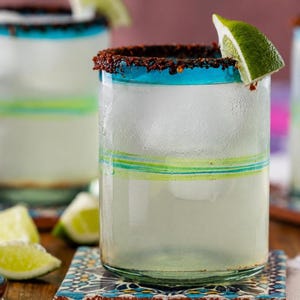 May include: A clear glass tumbler filled with ice and a pale liquid, featuring a dark red chili powder rim and a bright green lime wedge garnish. The glass has a thick teal blue rim and horizontal green and teal stripes around the middle section.