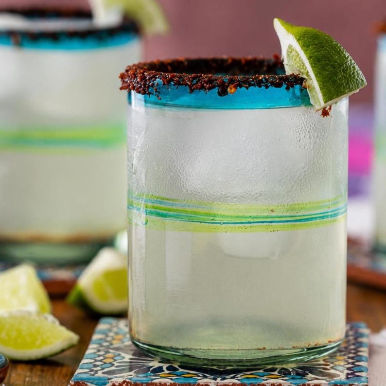 May include: A clear glass tumbler filled with ice and a pale liquid, featuring a dark red chili powder rim and a bright green lime wedge garnish. The glass has a thick teal blue rim and horizontal green and teal stripes around the middle section.