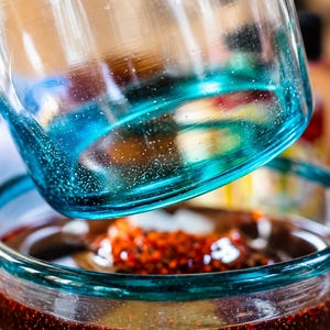 Puede incluir: Un recipiente de vidrio transparente con una base de degradado azul turquesa vibrante y una superficie texturizada. El recipiente est&aacute; colocado sobre un cuenco de vidrio a juego lleno de un l&iacute;quido marr&oacute;n rojizo oscuro con part&iacute;culas.