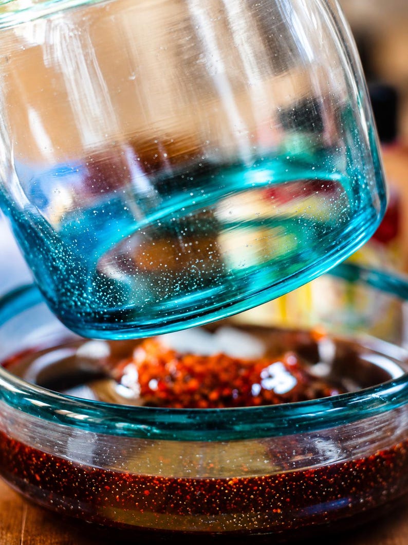 Puede incluir: Un recipiente de vidrio transparente con una base de degradado azul turquesa vibrante y una superficie texturizada. El recipiente est&aacute; colocado sobre un cuenco de vidrio a juego lleno de un l&iacute;quido marr&oacute;n rojizo oscuro con part&iacute;culas.