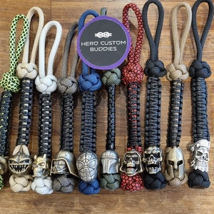 May include: A collection of 10 colourful paracord keychains with different metal charms. The charms include a shark, skull, Darth Vader, Spiderman, Mandalorian, skull, skull, a Spartan helmet, and a skull with a pirate hat. The keychains are all made with different coloured paracord and have a black and white pattern. The keychains are arranged in a row on a wooden surface. The text "HERO CUSTOM BUDDIES" is visible on a purple circle.
