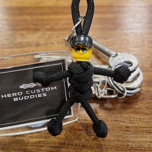 Puede incluir: Un llavero de paracord negro con una figura de Lego con gafas de sol y casco. La figura está unida a un cordón negro. Una etiqueta transparente con el texto "HERO CUSTOM BUDDIES" es visible.
