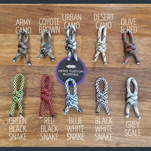 May include: A variety of paracord shoelace styles in different colors and patterns, including army camo, coyote brown, urban camo, desert camo, olive and red, green black snake, red black snake, blue white snake, black white snake, and grey scale. The shoelaces are tied in a knot and displayed on a wooden surface. The text "HERO CUSTOM BUDDIES" is visible in the center of the image.