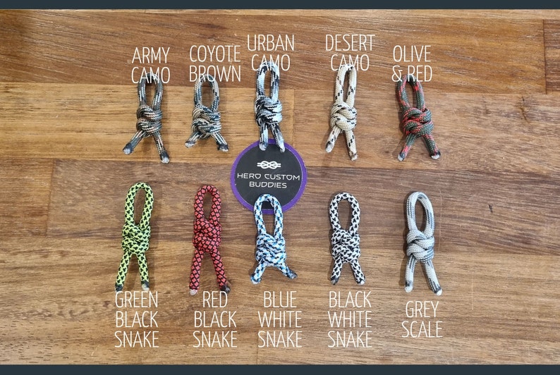 May include: A variety of paracord shoelace styles in different colors and patterns, including army camo, coyote brown, urban camo, desert camo, olive and red, green black snake, red black snake, blue white snake, black white snake, and grey scale. The shoelaces are tied in a knot and displayed on a wooden surface. The text "HERO CUSTOM BUDDIES" is visible in the center of the image.