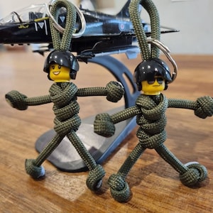 Puede incluir: Dos figuras de paracord verde con cabezas Lego negras y gafas de sol. Las figuras llevan atuendos de paracord verde y tienen cabezas Lego negras con gafas de sol. Las figuras están de pie sobre una superficie de madera.