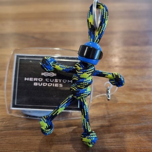 以下が含まれることがあります： 青いヘルメットと黒いバイザーをつけた青と緑のカラビナキーホルダーのフィギュア。フィギュアは腕を広げてポーズをとっています。キーホルダーは、「HERO CUSTOM BUDDIES」というテキストが書かれた透明なプラスチックカードに取り付けられています。