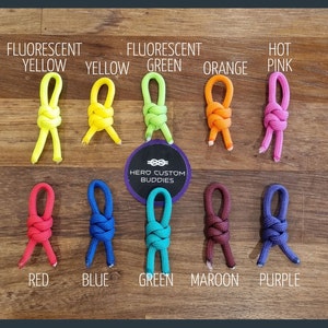 May include: A variety of colorful knotted cords in different colors, including fluorescent yellow, green, orange, pink, red, blue, green, maroon, and purple. The cords are arranged in two rows on a wooden surface. The text "HERO CUSTOM BUDDIES" is visible in the center of the image.
