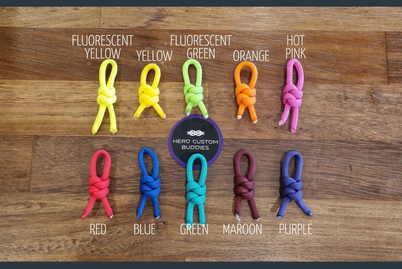 May include: A variety of colorful knotted cords in different colors, including fluorescent yellow, green, orange, pink, red, blue, green, maroon, and purple. The cords are arranged in two rows on a wooden surface. The text "HERO CUSTOM BUDDIES" is visible in the center of the image.